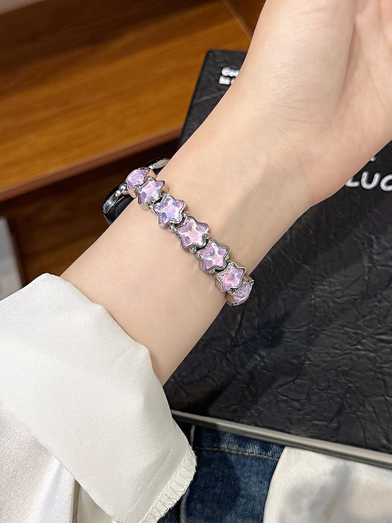 May include: A watch band featuring a series of linked, star-shaped charms with a pearlescent pink and purple finish. The star charms are connected by small silver-toned metal beads, creating a flexible bracelet design that attaches to a watch face.