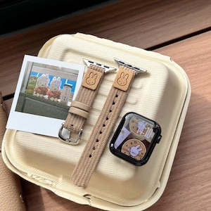 May include: A tan Apple Watch strap with a bunny motif, a black Apple Watch, and a photograph of two bunny figurines. The strap has a silver buckle and a woven texture. The watch face shows a bunny graphic.