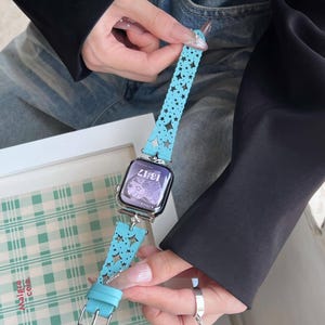 May include: A light blue watch band with a star pattern and a silver buckle. The watch face displays the time 13:17. The band is held by a person wearing a black jacket and blue jeans.