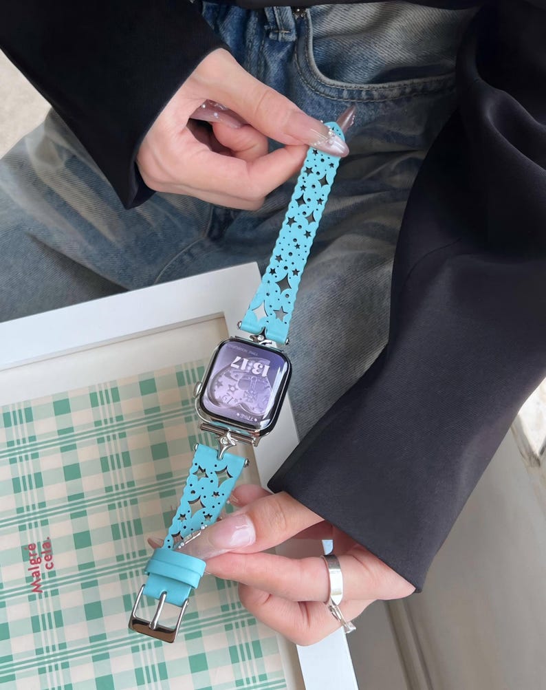 May include: A light blue watch band with a star pattern and a silver buckle. The watch face displays the time 13:17. The band is held by a person wearing a black jacket and blue jeans.