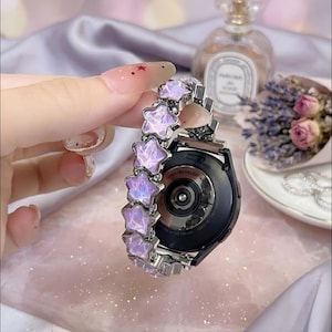 May include: A smartwatch band featuring a series of iridescent, light purple star-shaped links with a metallic silver finish. The band is attached to a black circular smartwatch casing, displaying the text Samsung Galaxy Watch on the back sensor.