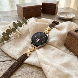 May include: A silver smartwatch with a brown leather band adorned with gold star-shaped accents and pearls. The watch face displays the time 2:28. The watch is set on a cream-colored cloth, with wooden boxes and a small dish in the background.