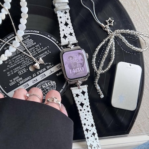 May include: A silver smartwatch with a white, star-patterned band and a lavender screen displaying the time. The image includes a matching beaded charm, a necklace with heart-shaped beads, and a silver bow pendant.