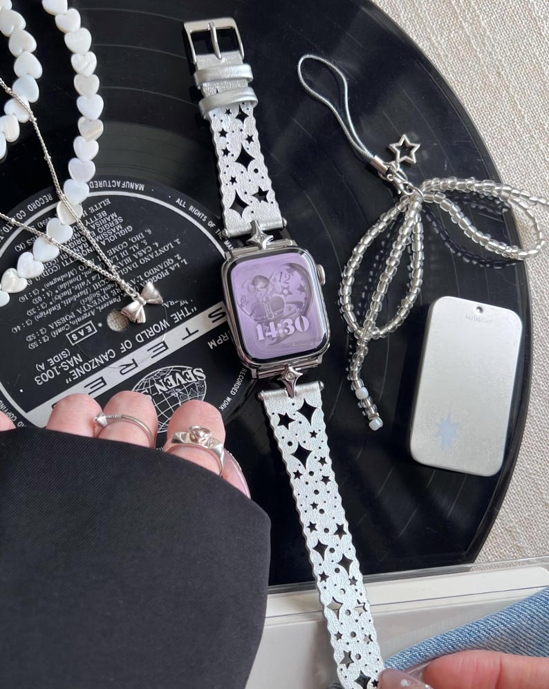 May include: A silver smartwatch with a white, star-patterned band and a lavender screen displaying the time. The image includes a matching beaded charm, a necklace with heart-shaped beads, and a silver bow pendant.