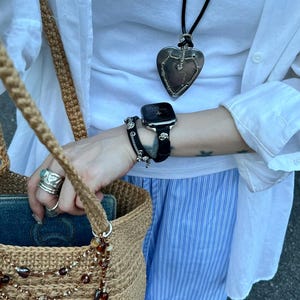 May include: A tan crocheted tote bag with a beaded strap, a silver heart pendant necklace, and a black smartwatch with a black band. The person is wearing a white shirt and blue and white striped pants.