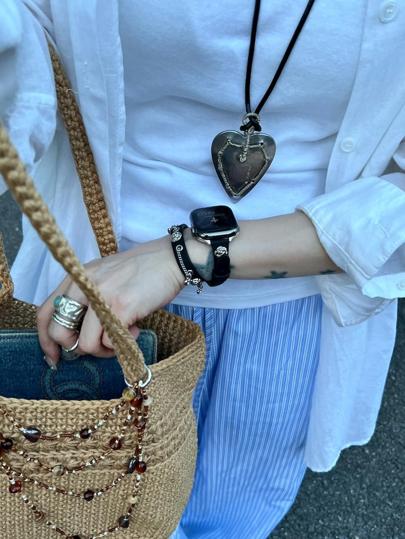 May include: A tan crocheted tote bag with a beaded strap, a silver heart pendant necklace, and a black smartwatch with a black band. The person is wearing a white shirt and blue and white striped pants.