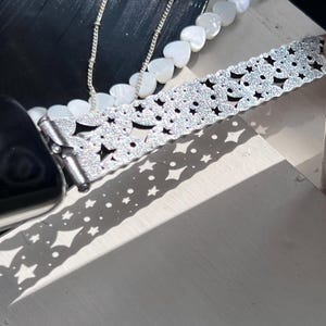 May include: A silver watch band with a star and floral pattern, sparkling with glitter. A silver chain necklace and a necklace with white heart-shaped beads are also visible. The items are on a white surface, with shadows.