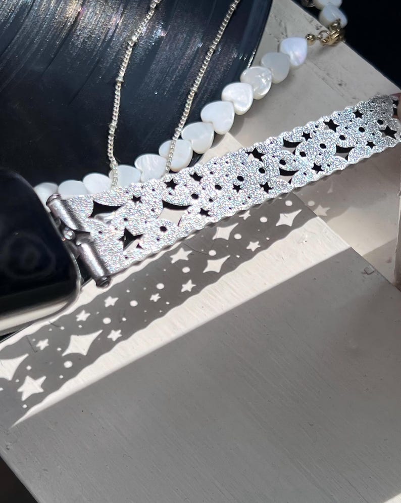 May include: A silver watch band with a star and floral pattern, sparkling with glitter. A silver chain necklace and a necklace with white heart-shaped beads are also visible. The items are on a white surface, with shadows.