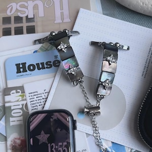 May include: A smartwatch with a black digital display showing stars and the time 13:55, attached to a decorative watch band featuring dark iridescent square shell tiles, silver-toned metal accents, clear crystal flower charms, and a dangling heart charm.