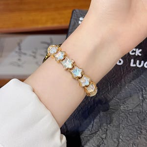 May include: A gold-toned bracelet featuring a series of connected star-shaped charms. Each star charm contains a translucent, iridescent center that reflects light with a pearlescent sheen. The bracelet is secured around the wrist with a metal link chain.