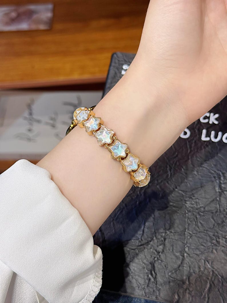 May include: A gold-toned bracelet featuring a series of connected star-shaped charms. Each star charm contains a translucent, iridescent center that reflects light with a pearlescent sheen. The bracelet is secured around the wrist with a metal link chain.