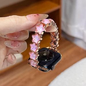 May include: A smartwatch band featuring a series of connected rose gold metal star-shaped links, each inlaid with a translucent, iridescent pink stone. The band is attached to a black smartwatch casing with a circular sensor on the back.