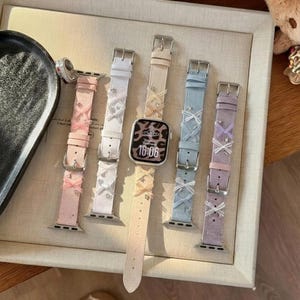 Könnte beinhalten: Eine Auswahl an pastellfarbenen Uhrenarmbändern mit silbernen Schnallen und dekorativen Schleifen. Die Armbänder sind in Rosa, Weiß, Beige, Blau und Lavendel gehalten. Eine Smartwatch mit Leopardenmuster-Display zeigt 16:06 an.