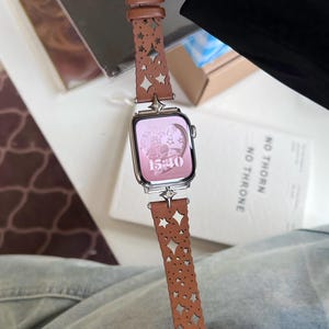 May include: A silver smartwatch with a pink screen displaying the time 15:40. The watch has a brown leather band with star and geometric cutouts. The band is attached with silver hardware.
