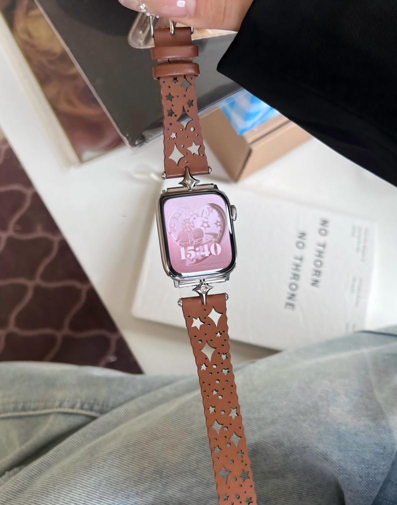 May include: A silver smartwatch with a pink screen displaying the time 15:40. The watch has a brown leather band with star and geometric cutouts. The band is attached with silver hardware.