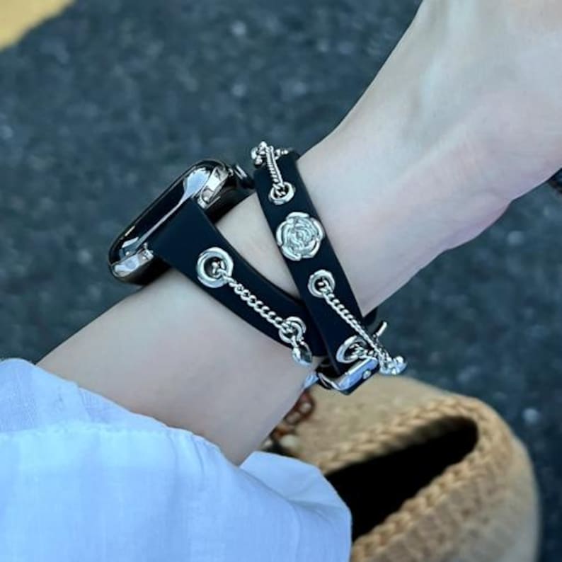 May include: Black leather watch band with silver-tone chain and rose accents. The watch band is attached to a silver watch. The watch band is worn on a wrist.
