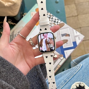 May include: A white smartwatch with a band featuring black heart-shaped patterns. The watch face displays a selfie with the time 1:58. A small charm hangs from the band. The watch is held by a person wearing rings.
