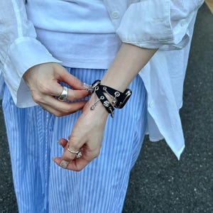 May include: Black leather bracelet with silver-tone studs and chain details. The bracelet is worn on a wrist, and the person is wearing a white shirt and blue striped pants. The bracelet has a gothic or punk aesthetic.