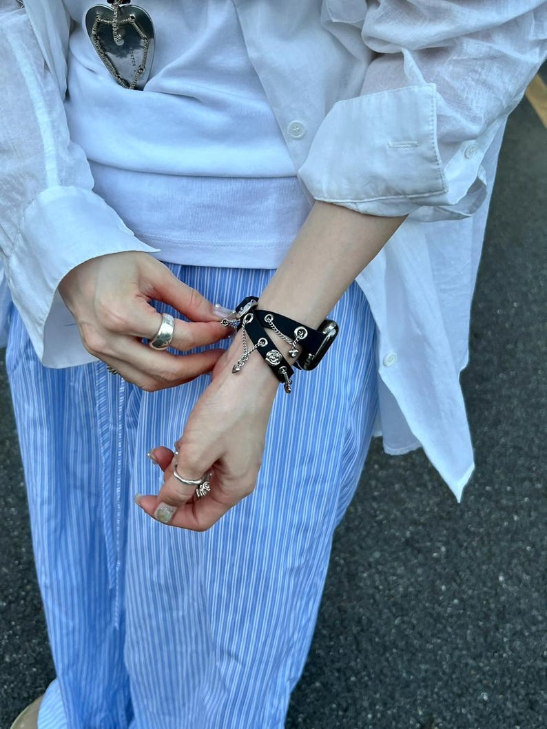 May include: Black leather bracelet with silver-tone studs and chain details. The bracelet is worn on a wrist, and the person is wearing a white shirt and blue striped pants. The bracelet has a gothic or punk aesthetic.