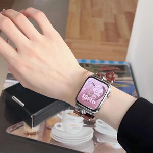 May include: A silver smartwatch with a brown leather band is displayed on a wrist. The watch face is pink and shows the time 15:46, with decorative elements. The band has a decorative design.