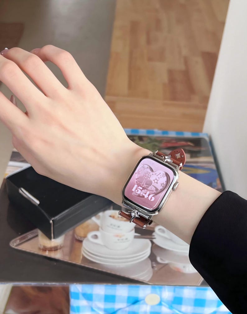 May include: A silver smartwatch with a brown leather band is displayed on a wrist. The watch face is pink and shows the time 15:46, with decorative elements. The band has a decorative design.