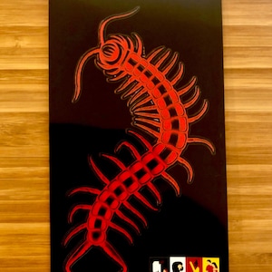 May include: A red centipede illustration on a black background. The centipede is stylized with a detailed, segmented body and multiple legs. The image has a glossy finish and a white border. A small, colorful graphic is at the bottom.