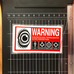 Könnte beinhalten: Ein rot-weißer Warnaufkleber mit einer Augengrafik und dem Text "WARNING CONTENTS MAY PERMANENTLY ALTER PERCEPTION". Der Aufkleber enthält auch Symbole für Leben, Strategie, Richtung und Ziel. Auf einem schwarzen Papierschneider.
