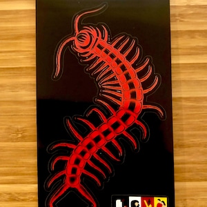 May include: A black sticker featuring a red centipede design. The centipede is stylized with a detailed, segmented body and multiple legs. A small, colorful graphic of people is in the lower right corner. The sticker is on a white background.