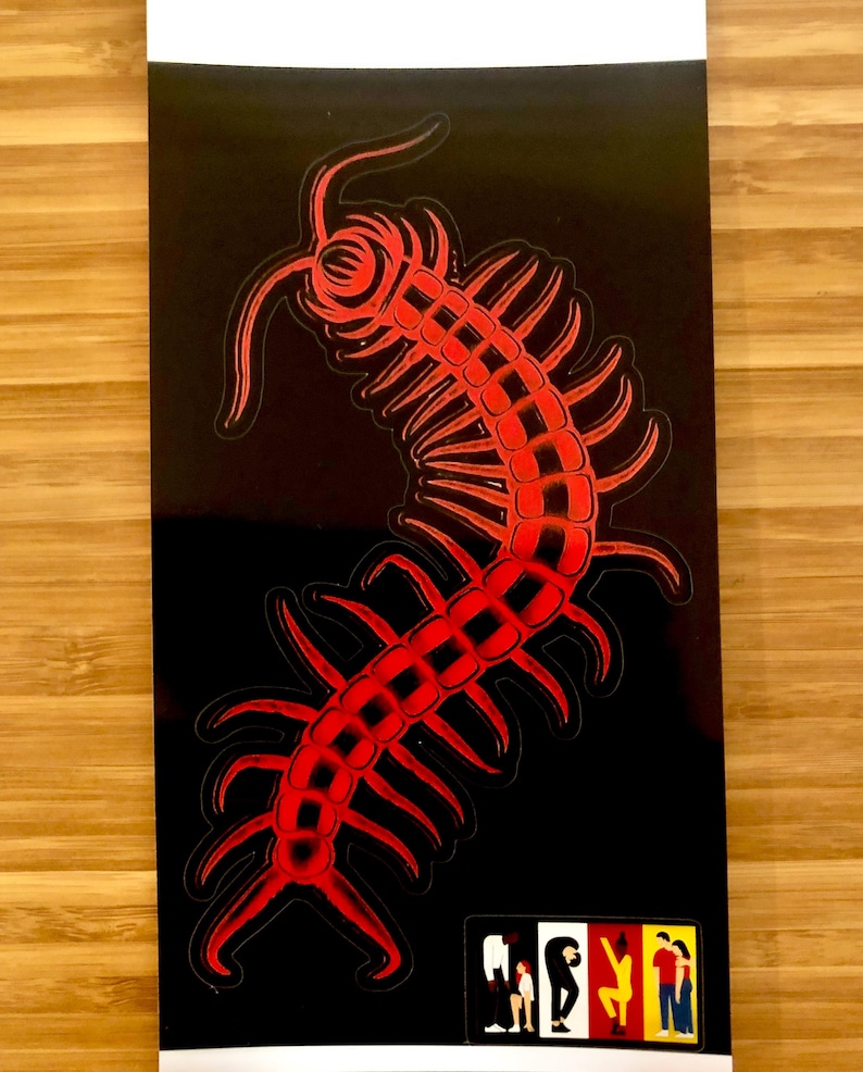 May include: A black sticker featuring a red centipede design. The centipede is stylized with a detailed, segmented body and multiple legs. A small, colorful graphic of people is in the lower right corner. The sticker is on a white background.