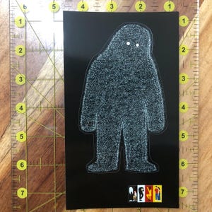 May include: A black sticker featuring a stylized, glittery figure with two white eyes. The figure is standing upright. A small, colorful logo is at the bottom. The sticker is placed on a wooden surface next to a ruler.