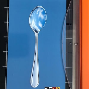 Può includere: Un cucchiaio in acciaio inossidabile con un riflesso del cielo nuvoloso su sfondo blu. Il cucchiaio è posizionato su carta blu, con una piccola immagine di persone nell'angolo in basso a destra. L'immagine è su un tappetino da taglio nero.