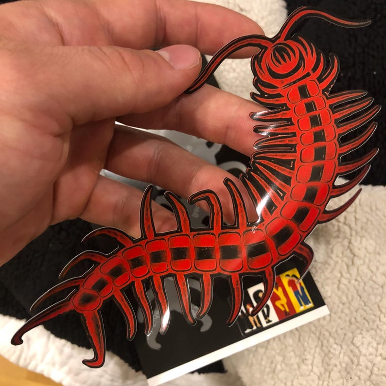 May include: A red and black centipede sticker with a glossy finish. The sticker is held up by a hand, and the background is a black and white patterned surface. The centipede has a detailed design.