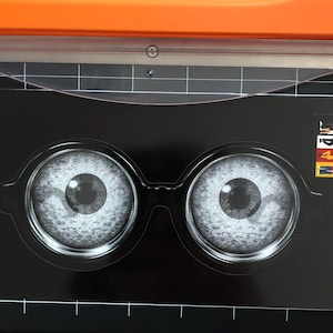 May include: A black graphic with two large, detailed eye illustrations, resembling eyeglasses. The eyes are light gray with dark pupils. The graphic is on a black surface with a ruler and orange cutting tool in the background.
