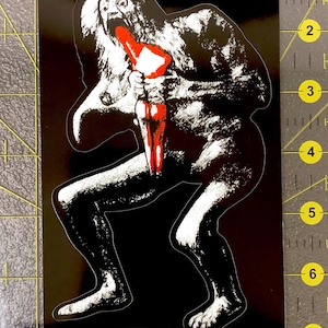 Puede incluir: Pegatina negra con una ilustración blanca y roja de una figura con la boca abierta y un objeto rojo. La pegatina tiene un pequeño diseño rectangular en la parte inferior con figuras estilizadas.