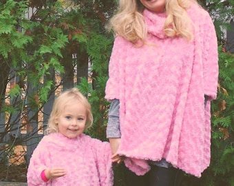 Peut inclure: Deux personnes portant des ponchos en fourrure rose. L'adulte porte un legging noir et des ballerines noires. L'enfant porte un legging noir et des baskets bleues. Elles sont debout devant un buisson vert et un mur en pierre.
