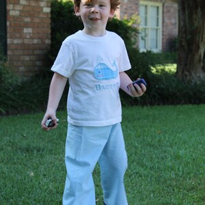 May include: A young boy wearing a white t-shirt with a blue whale design and the word "Barrie" embroidered on it. He is wearing light blue pants and is standing on a grassy lawn.