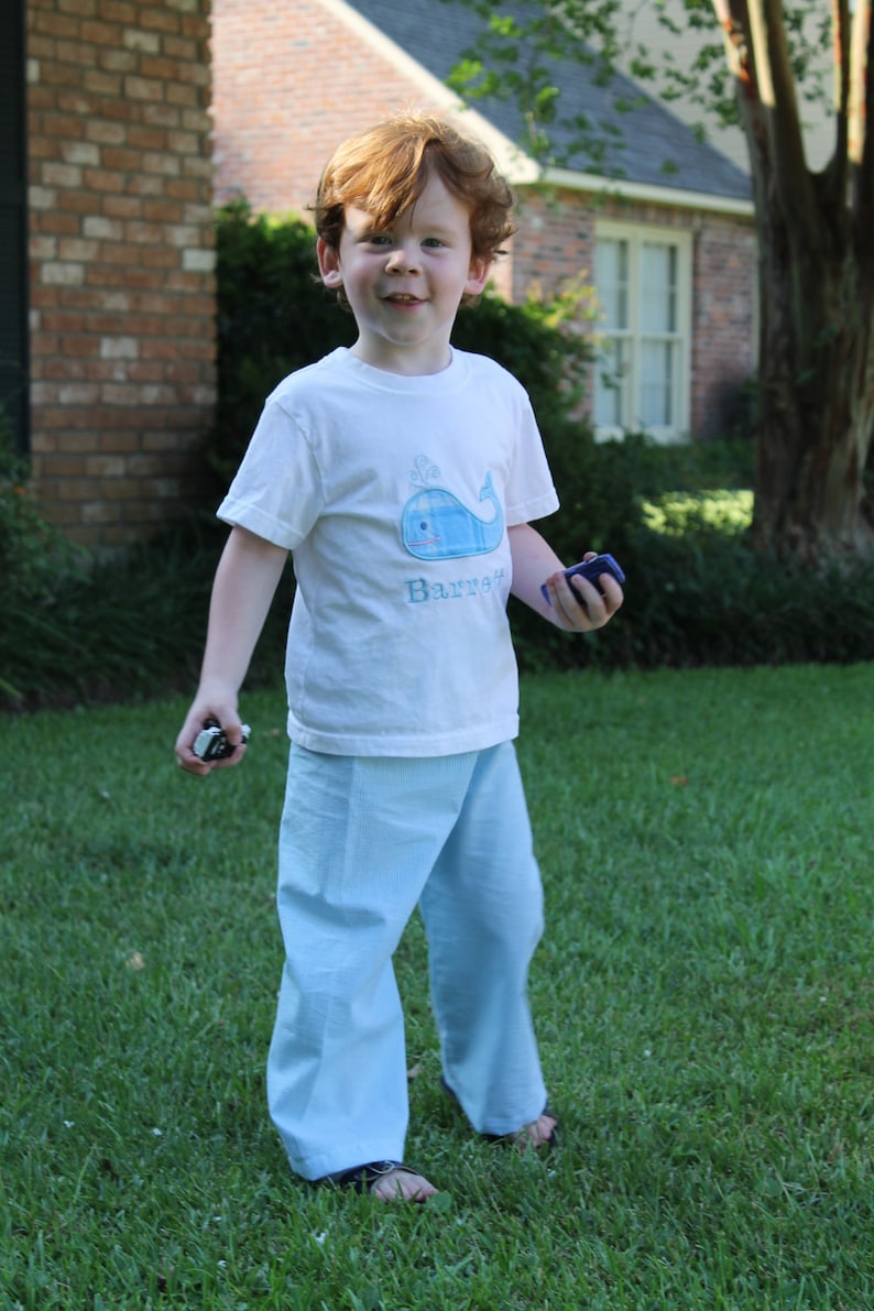 May include: A young boy wearing a white t-shirt with a blue whale design and the word "Barrie" embroidered on it. He is wearing light blue pants and is standing on a grassy lawn.