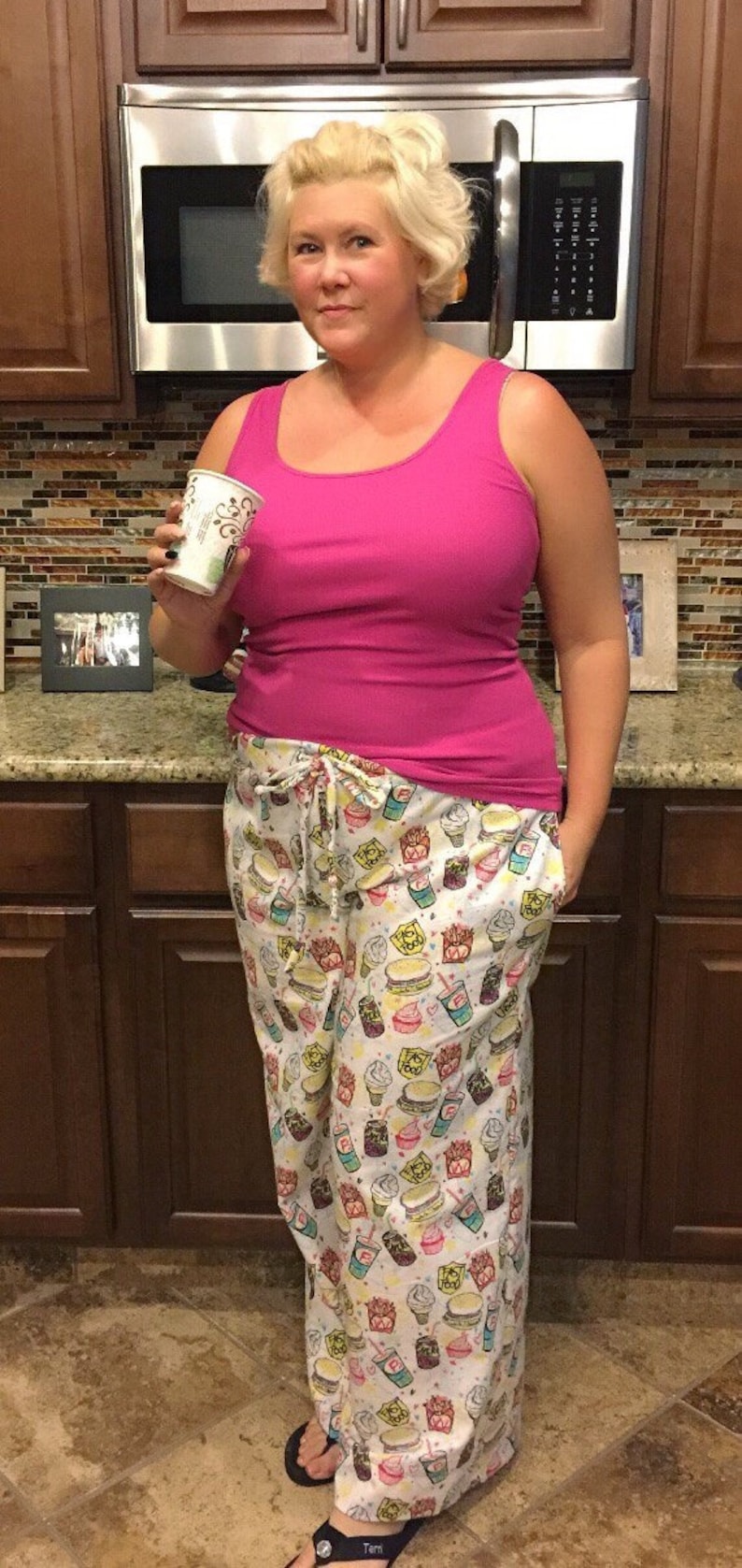 May include: A woman wearing a pink tank top and white pajama pants with a food print. The pants have a drawstring waist and a variety of food items printed on them, including ice cream cones, hamburgers, french fries, and soda.
