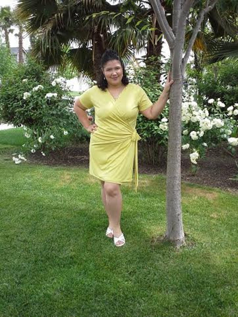 May include: A woman wearing a yellow wrap dress and white sandals stands in a garden with green grass and white flowers.