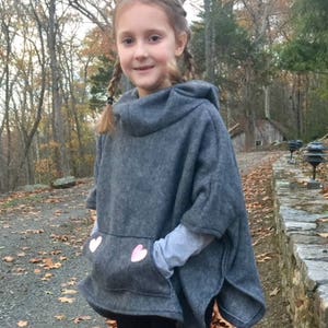 Könnte beinhalten: Ein grauer Kapuzenponcho mit rosa Herz-Applikationen auf der Tasche. Der Poncho wird von einem jungen Mädchen mit geflochtenen Haaren getragen.
