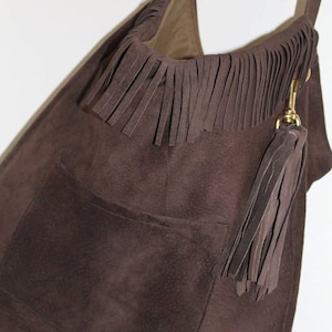 Puede incluir: Un bolso de mano de ante marrón con ribete de flecos y un borla. El bolso tiene un bolsillo grande en la parte delantera.