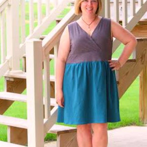 May include: A woman wearing a sleeveless dress with a gray top and a teal blue bottom. The dress has a wrap style bodice and a gathered skirt. She is standing on a wooden staircase with white railings.