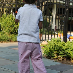May include: A young boy wearing a white short-sleeved shirt and pink and white striped pants with a sailboat pattern. He is standing on a stone patio.
