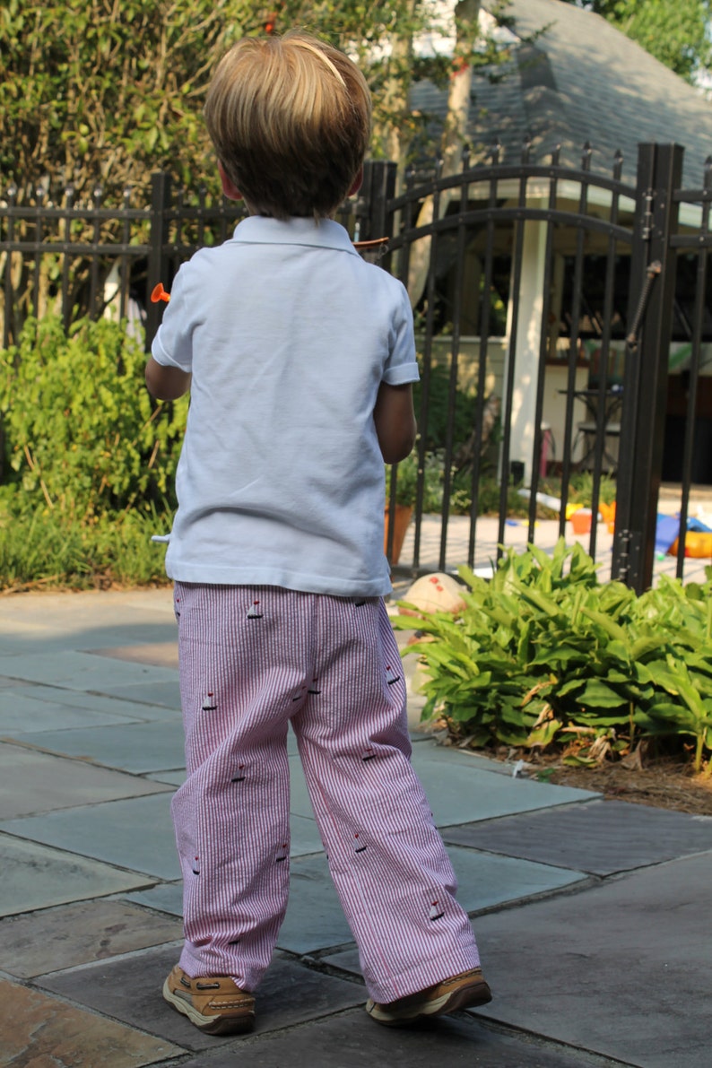 May include: A young boy wearing a white short-sleeved shirt and pink and white striped pants with a sailboat pattern. He is standing on a stone patio.