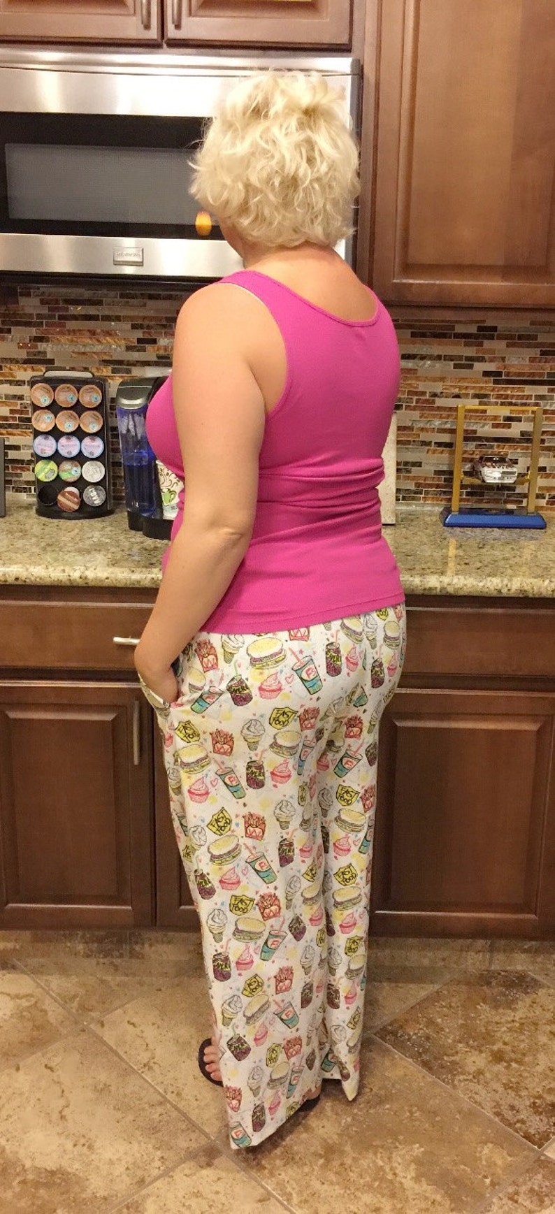 May include: A woman wearing a pink tank top and white pajama pants with a fast food print. The pants feature images of hamburgers, french fries, ice cream cones, and soda cups.