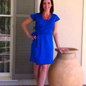 May include: A woman wearing a bright blue wrap dress with short sleeves and an orange beaded necklace. She is standing in front of a large terracotta pot.