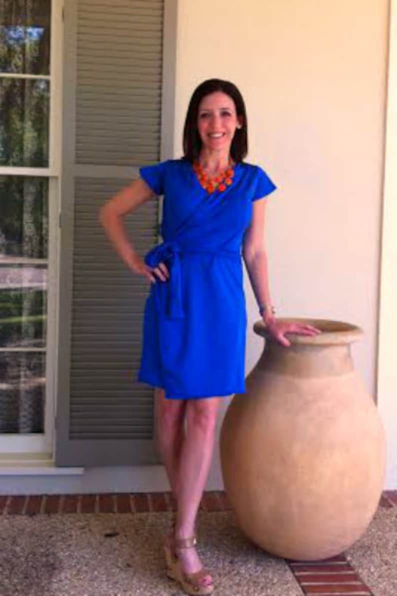May include: A woman wearing a bright blue wrap dress with short sleeves and an orange beaded necklace. She is standing in front of a large terracotta pot.