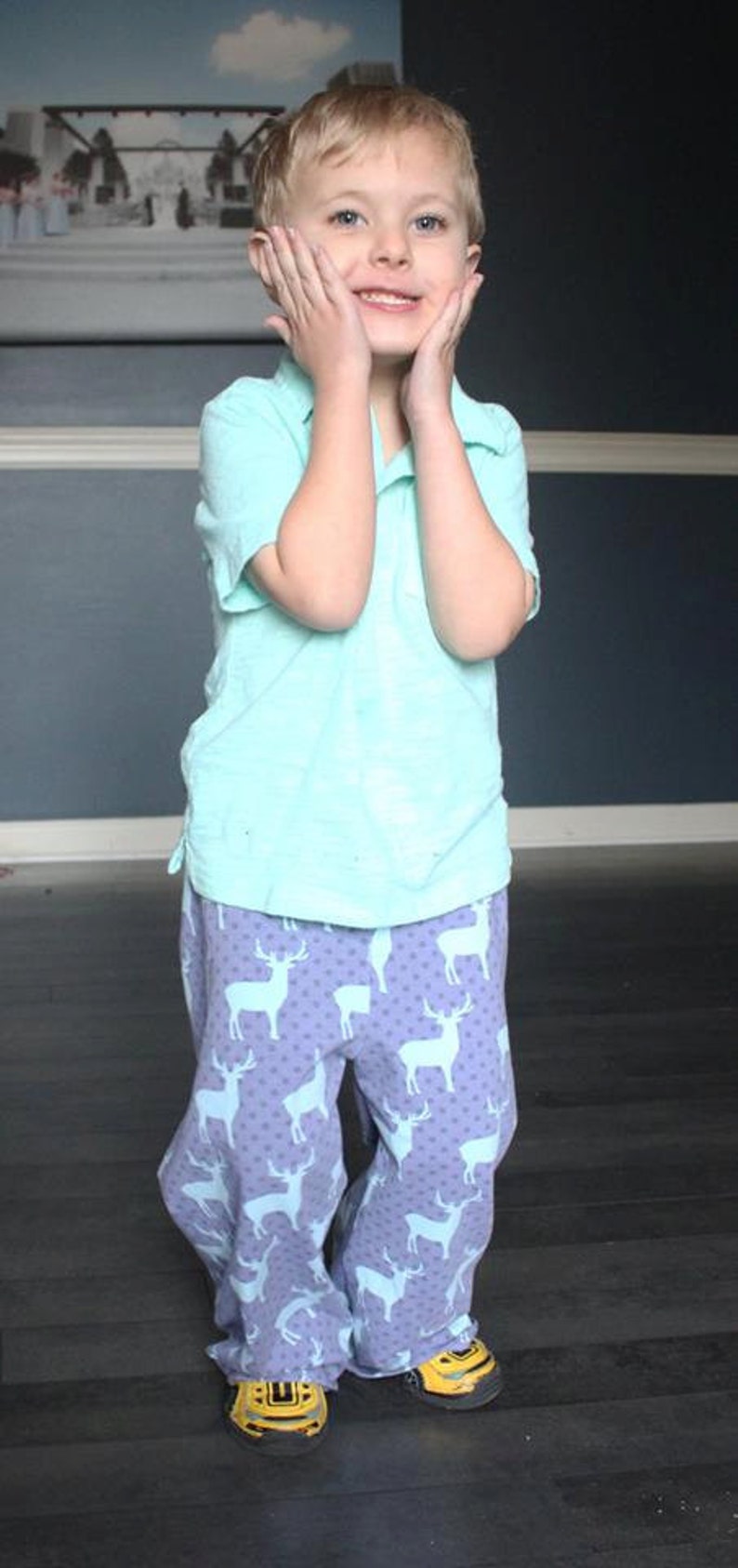 May include: A young boy wearing a light blue polo shirt and purple pants with a white deer pattern. He is smiling and has his hands on his cheeks.