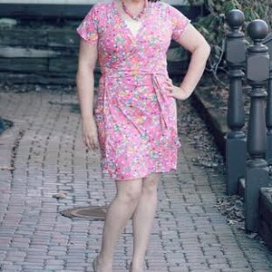 May include: A woman wearing a pink floral wrap dress with a white top underneath. She is standing on a brick pathway.