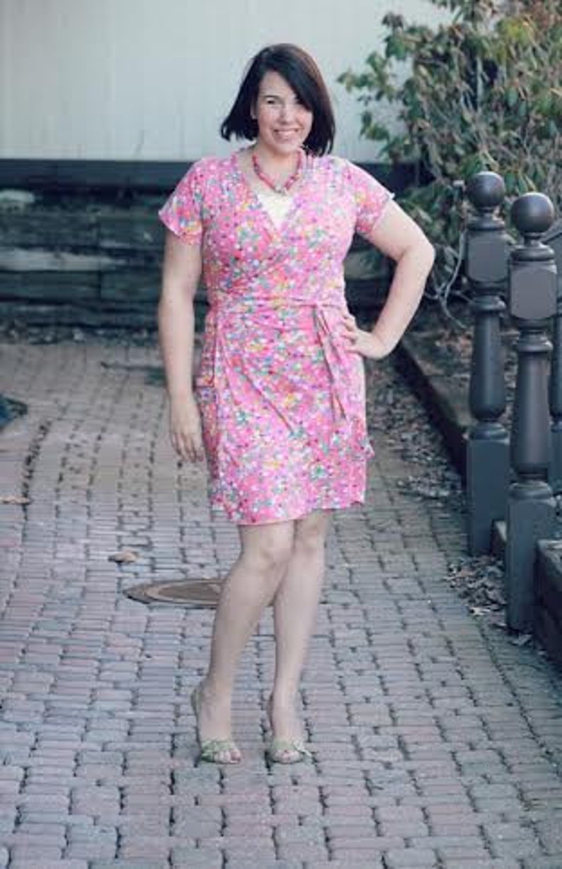 May include: A woman wearing a pink floral wrap dress with a white top underneath. She is standing on a brick pathway.
